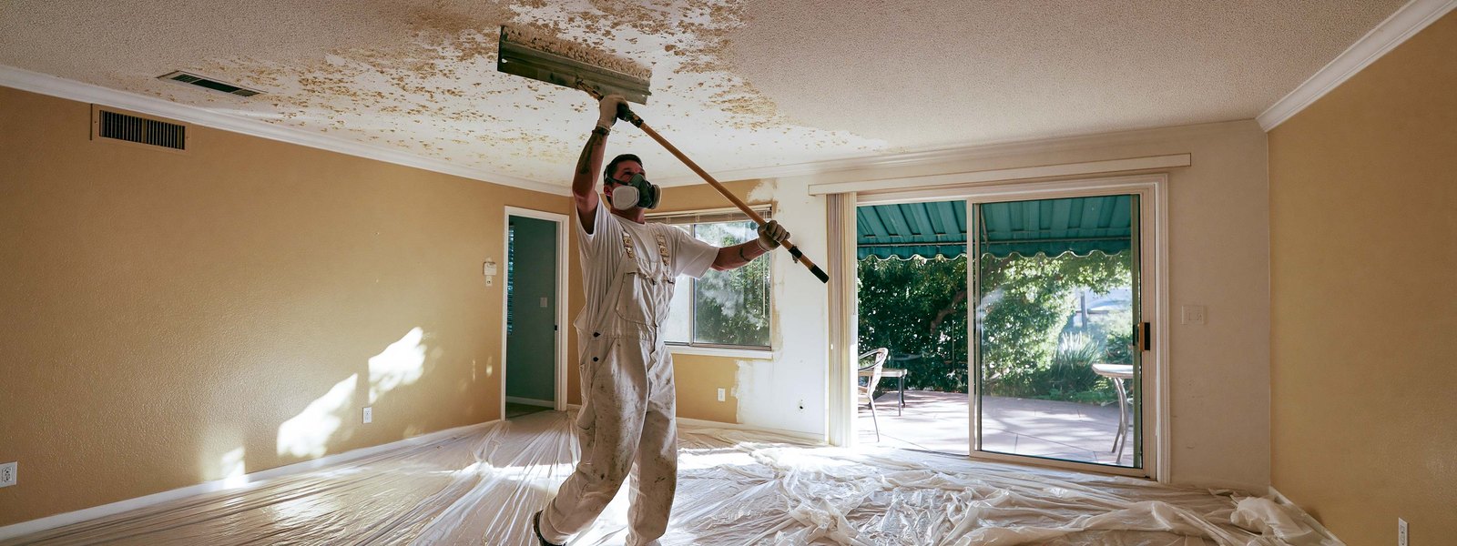 A contractor in a respirator scraping a sprayed ceiling, the floor below covered in sheeting