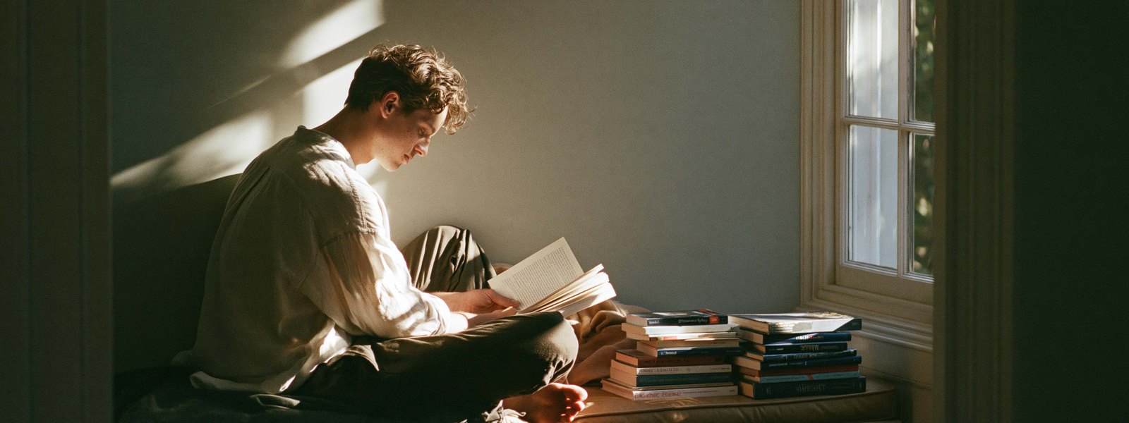A young man reading in a sunlit window seat with a stack of books beside him